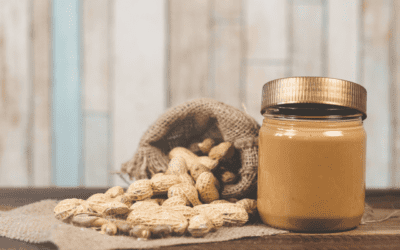 A burlap bag of unshelled peanuts spilling over on to a table next to a jar of peanut butter.