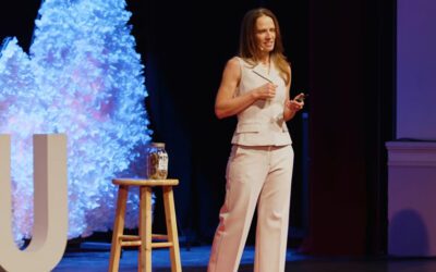 Aimee Bernard, PhD, stands on stage for a TEDx Talk, next to a swear jar labelled "MOM" on a stool.