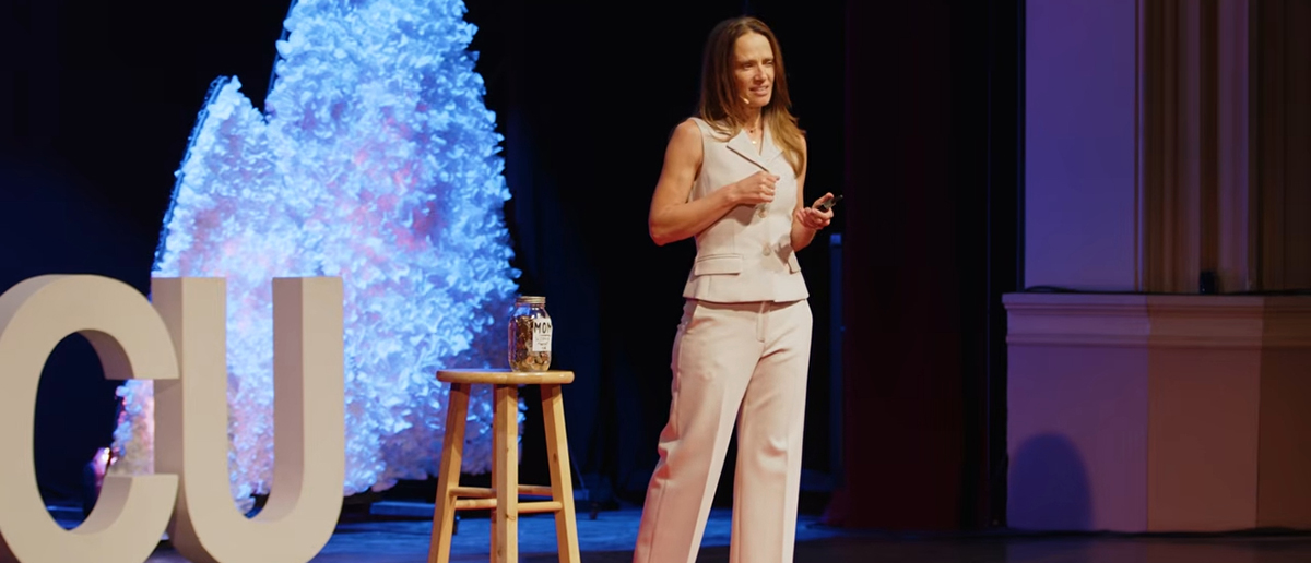 Aimee Bernard, PhD, stands on stage for a TEDx Talk, next to a swear jar labelled "MOM" on a stool.